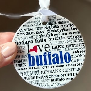 May include: White ceramic ornament with a red and blue outline of New York State and the word "love" in front of the state. The ornament is covered in black text that lists various landmarks, businesses, and attractions in Buffalo, New York.