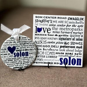 May include: A white ceramic ornament with a blue heart and the word "Solon" in black text. The ornament is surrounded by a word cloud of local businesses and landmarks in Solon, Ohio. The word cloud includes names like "Stouffer's", "Quinn's", "Jim's", "Alescis", "Chicago Deli", "Solon Hardware", "Solon Comets", "Carter Lumber", "Pettibone Rd", and "Grantwood Golf Course".