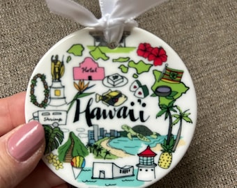 Puede incluir: Adorno de cerámica blanca con una ilustración colorida de lugares emblemáticos de Hawái y la palabra "Hawaii" en escritura negra. El adorno tiene una cinta blanca para colgar.