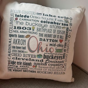 May include: Square off-white pillow with the word "Ohio" in the center, surrounded by text and icons representing Ohio landmarks and attractions. The pillow features a variety of fonts and colors.