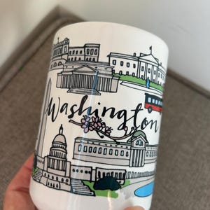 May include: White ceramic mug featuring a black and white illustrated design of Washington D.C. landmarks, including the Capitol Building and the White House. The word "Washington" is written in black script. A red bus is also depicted.