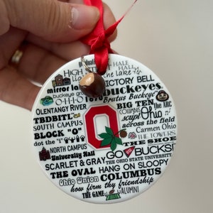 May include: White ceramic ornament with a red and white Ohio State University logo. The ornament is covered in text, including "The Oval", "Go Bucks", "Scarlet & Gray", "Hang on Sloopy", "Ohio Union", "Columbus", and "How firm thy friendship".
