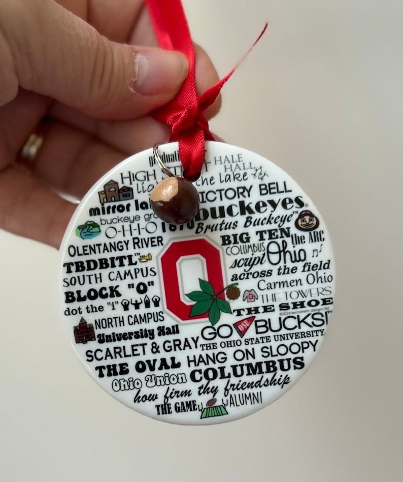 May include: White ceramic ornament with a red and white Ohio State University logo. The ornament is covered in text, including "The Oval", "Go Bucks", "Scarlet & Gray", "Hang on Sloopy", "Ohio Union", "Columbus", and "How firm thy friendship".