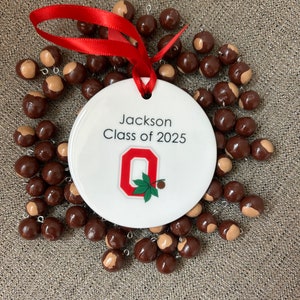 May include: White ceramic ornament with a red ribbon, featuring the text "Jackson Class of 2025" and a red "O" with a green buckeye leaf. The ornament is surrounded by brown buckeye nuts.