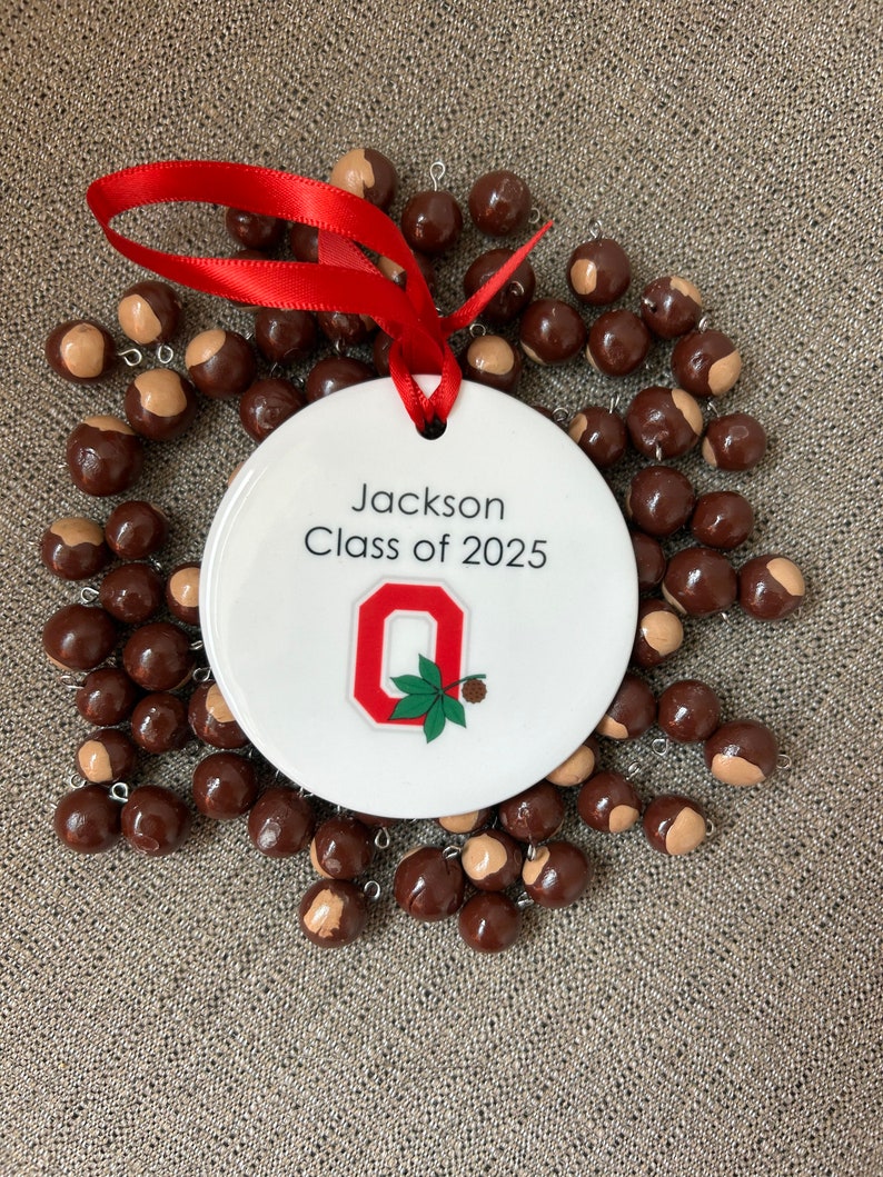 May include: White ceramic ornament with a red ribbon, featuring the text "Jackson Class of 2025" and a red "O" with a green buckeye leaf. The ornament is surrounded by brown buckeye nuts.