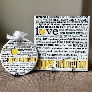 May include: A white ceramic tile with black text listing places and landmarks in Upper Arlington, Ohio. The text is arranged in a circular pattern around a yellow outline of the state of Ohio. The tile is on a stand.