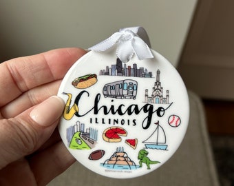 Pode incluir: Ornamento de cerâmica branca com uma ilustração colorida de marcos de Chicago, incluindo o horizonte, um cachorro-quente, uma pizza, um veleiro, uma bola de beisebol, uma bola de futebol, um dinossauro e as palavras "Chicago Illinois".