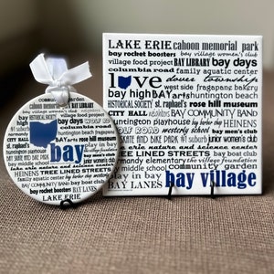 May include: A white ceramic ornament and a tile with the words "bay" and "bay village" in blue. The ornament has a blue outline of a state and the words "Lake Erie" and "bay" in blue. The tile has a list of words.