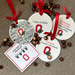 May include: Four white ceramic ornaments with red ribbon and a red and white Ohio State University logo. The ornaments feature the text "Class of 2024", "Ella Modine Class of 2025 Bachelor of Science Accounting", "Charlotte Forever a Buckeye!" and "How Firm Thy Friendship". The ornaments are surrounded by brown buckeye nuts.
