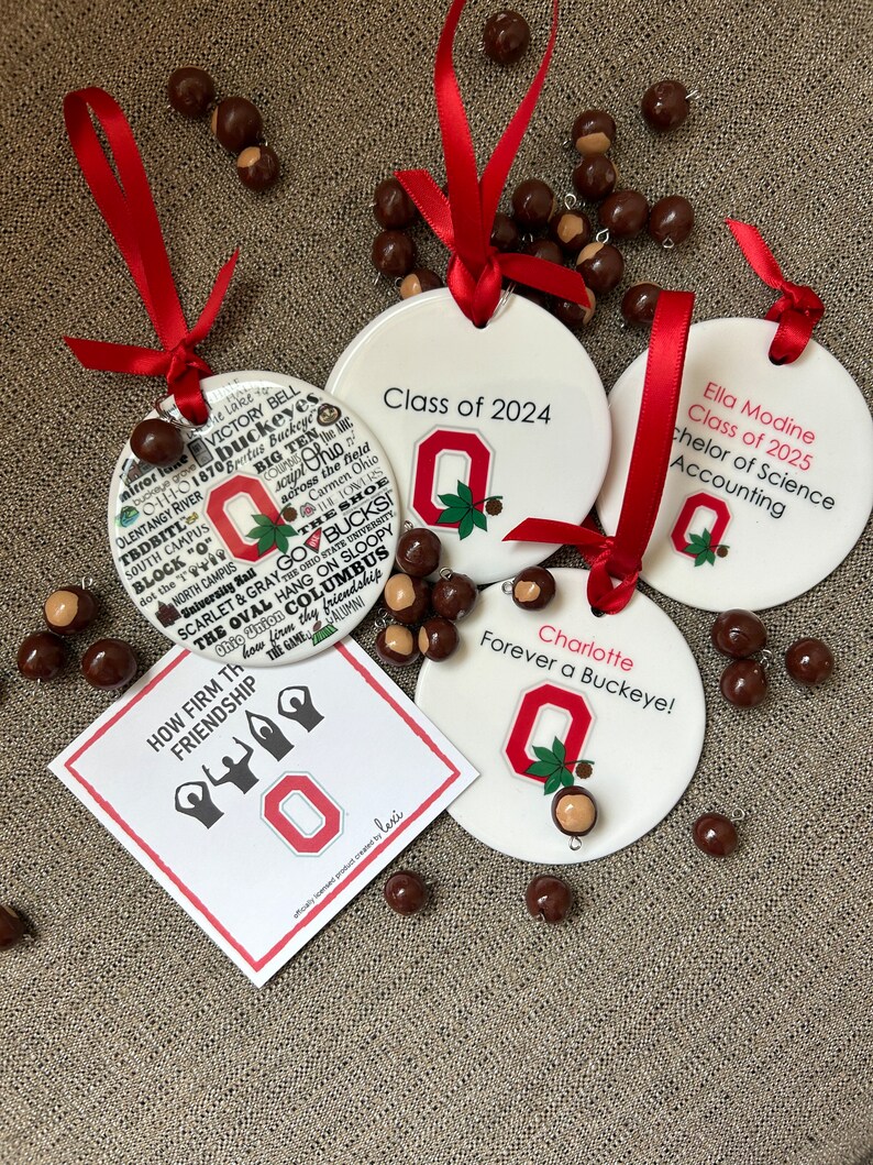 May include: Four white ceramic ornaments with red ribbon and a red and white Ohio State University logo. The ornaments feature the text "Class of 2024", "Ella Modine Class of 2025 Bachelor of Science Accounting", "Charlotte Forever a Buckeye!" and "How Firm Thy Friendship". The ornaments are surrounded by brown buckeye nuts.