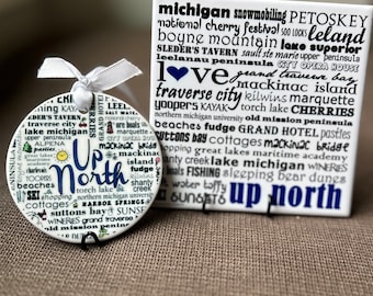 May include: A white ceramic ornament with the words "Up North" in blue lettering. The ornament is decorated with various words and phrases related to Michigan, including "traverse city", "Mackinac Island", "Sleeping Bear Dunes", and "Lake Superior".
