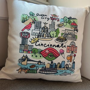 May include: Cream-colored throw pillow with a colorful Cincinnati, Ohio, design. The illustration includes landmarks, a baseball stadium, and the words "Queen City" and "Cincinnati".