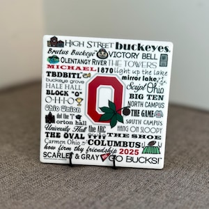 May include: White ceramic tile with a red and black Ohio State University logo and a collage of words and phrases related to the university, including "Buckeyes", "Victory Bell", "The Oval", "Script Ohio", "The Game", and "Go Bucksi!"