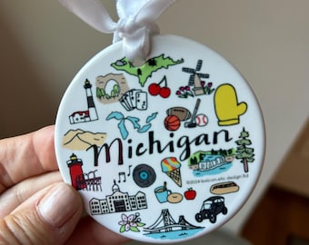 May include: White ceramic ornament with a colorful illustration of Michigan landmarks and the word "Michigan". The ornament features a lighthouse, a windmill, a mitten, a cherry, a bridge, a hot dog, and other iconic symbols of the state.