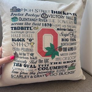 May include: A beige square pillow with a printed design featuring the Ohio State University logo and various related words and phrases. The design includes the words "High Street Buckeyes," "Script Ohio," and "Go Bucks!"