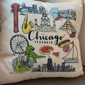 May include: A cream-colored decorative pillow featuring a colorful Chicago, Illinois themed design. The design includes a Ferris wheel, saxophone, dinosaur, pizza, and the Chicago skyline. The word "Chicago" is written in a black script font.