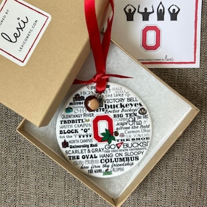 May include: A white round ceramic ornament with a red and white Ohio State University logo in the center. The ornament is covered in white text that includes the words "Go Bucks", "The Shoe", "Scarlet & Gray", "The Oval", "Hang on Sloopy", and "Columbus".