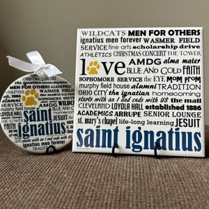 May include: Two white ceramic tiles with blue and gold lettering. The tile on the left has a paw print with the words "Saint Ignatius" and other text. The tile on the right has the words "Wildcats Men for Others" and other text.