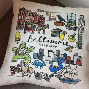 May include: A decorative pillow with a Baltimore, Maryland themed design. The pillow features colorful illustrations of local landmarks, a horse-drawn carriage, a crab, and the city skyline. The word "Baltimore" is prominently displayed.
