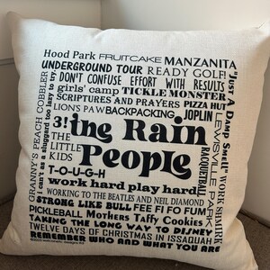 May include: Cream-colored square pillow with black text. The words "The Rain People" are prominently displayed, surrounded by phrases like "Work Hard Play Hard" and "Twelve Days of Christmas." A decorative accent for any room.
