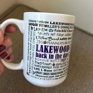 May include: White ceramic mug featuring the text "Lakewood back in the day" in purple, with various local businesses and landmarks listed in black. The mug has a curved handle and is held by a person.