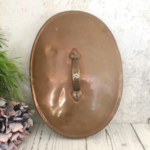 Puede incluir: Tapa de olla ovalada de cobre con asa. La tapa tiene una superficie brillante y reflectante y un asa con el texto "JPD. 6.5". El asa está sujeta con remaches. La tapa es de color cobre cálido.