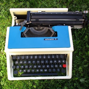 May include: A white and blue Olivetti T typewriter with a black keyboard and a red button. The typewriter is sitting on a bed of green grass.