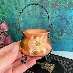 May include: A small copper-colored decorative pot with a black twisted handle and legs. The pot features a painted white, yellow, and blue floral design on the front. The pot is held in a person's hand against a colorful background.