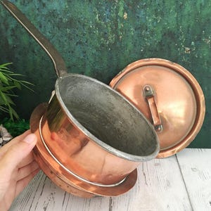 Könnte beinhalten: Kupfer-Kochtopf mit langem Griff und passendem Deckel. Das Innere des Topfes ist von stumpfer silberner Farbe und kontrastiert mit dem glänzenden Kupferäußeren. Der Deckel hat einen kleinen Griff.