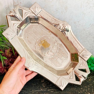 Puede incluir: Bandeja plateada de estilo Art Déco con un centro rectangular y detalles geométricos. La bandeja tiene un borde decorativo con formas florales y geométricas estilizadas. La bandeja está sujeta por una mano.