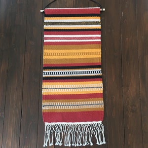 Op de afbeelding: Een geweven wandkleed met een gestreept patroon in rood, goud, zwart, wit en geel. Het rechthoekige textiel heeft een franje aan de onderkant en is opgehangen aan een houten deuvel met een zwart koord.