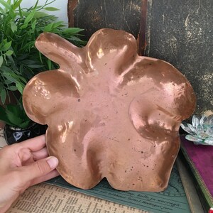 May include: A polished copper dish shaped like a five-petaled flower. The metal surface features a hammered texture with subtle indentations and a warm, metallic reddish-brown tone. The dish has a smooth, reflective finish with visible natural patina.
