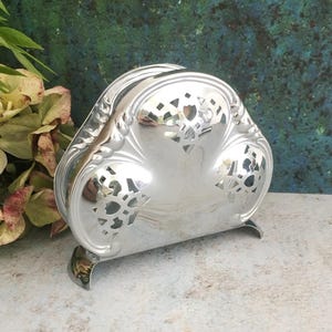 May include: A silver-colored napkin holder with a decorative floral design. The holder has a curved shape with cut-out patterns and rests on small feet. The polished surface reflects the surrounding environment.