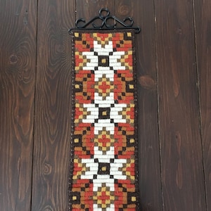 Könnte beinhalten: Ein Vintage-Wandbehang mit einem geometrischen Muster in Braun-, Orange-, Weiß- und Gelbtönen. Der Behang ist aus Garn gefertigt und an einem schwarzen Metallhalter befestigt.