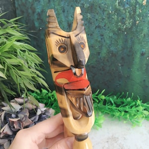 Op de afbeelding: Een gesneden houten totemfiguur met een gestileerd gezicht, bruine en zwarte details, rode accenten en een gestreept patroon. De figuur heeft een lang, verticaal ontwerp en wordt tegen een groen-blauwe achtergrond gehouden.