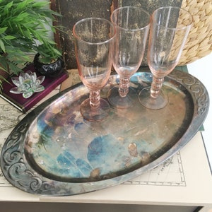 Peut inclure: Trois flûtes en verre rose clair avec des pieds texturés sont posées sur un plateau ovale en métal argenté terni. Le plateau présente une bordure ornementale en relief et une surface patinée et usée aux tons bleus et marron.