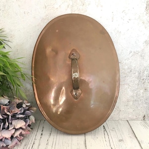 Puede incluir: Tapa ovalada de olla de cobre con asa de bronce. La tapa tiene una superficie ligeramente curvada y un asa de bronce sujeta con remaches. El asa tiene grabado el texto "JPG 6.5". El cobre tiene un color marrón rojizo cálido.