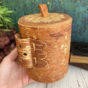 May include: A cylindrical birch bark box with a leather lid and strap. The box is a natural brown colour with a light brown leather strap and lid.