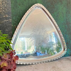 May include: A silver-colored, triangular serving tray with a decorative, etched design in the center. The tray has a scalloped edge and reflects the surrounding environment. The tray is likely made of metal.