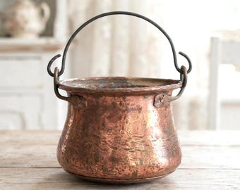 Puede incluir: Una olla redonda de cobre martillado con un asa giratoria de metal oscuro sujeta a dos bucles laterales. La superficie exterior presenta un acabado texturizado y desgastado con tonos de cobre metálico y marrón, mostrando signos de uso y antigüedad.