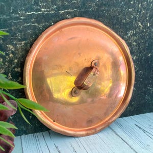 Puede incluir: Una tapa de olla redonda de cobre con asa. La tapa tiene un borde ligeramente festoneado y un acabado deslustrado. El asa está sujeta con dos remaches y tiene forma rectangular. La tapa es probablemente vintage y muestra signos de uso.