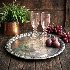 May include: A round, hammered silver-toned metal tray with a scalloped edge, two tall pink-tinted glass goblets with spherical stems, two fresh purple figs, and a cluster of red grapes, all arranged together on a dark wooden surface.