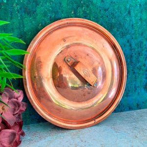 May include: A round copper pot lid with a handle. The lid has a shiny, reflective surface and a slightly raised rim. The handle is rectangular and attached to the lid with a small hinge. The lid is set against a teal and blue background.