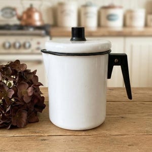 Könnte beinhalten: Eine weiße Emaille-Kaffeekanne mit schwarzem Rand, schwarzem Deckelknopf und schwarzem Griff. Das zylindrische Gefäß hat eine glatte Oberfläche und ein schlichtes, funktionales Design. Sie steht neben einem Strauß getrockneter brauner Hortensien.