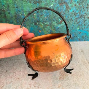 May include: A small, hammered copper cauldron with a black handle and decorative feet. The cauldron has a shiny, reflective surface and is designed with a rounded shape. The handle is twisted, and the feet are stylized.