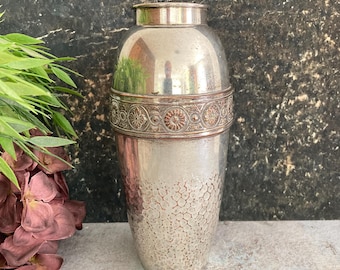 May include: A vintage silver cocktail shaker with a hammered texture on the lower portion. The shaker features a decorative band with floral and circular patterns. The top and base are smooth and shiny, reflecting the surrounding environment.