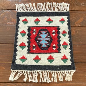 May include: A small, rectangular woven rug with a central red and blue design. The rug features a black border and white fringe. The pattern includes red and green floral motifs on a cream background.