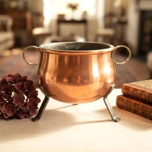 Op de afbeelding: Een gepolijste koperen pot met een rond lichaam, twee handvatten aan de zijkanten en drie schuine metalen poten. De pot heeft een glad, reflecterend oppervlak en een brede, cirkelvormige opening aan de bovenzijde.