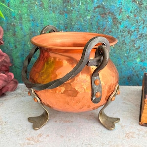 Op de afbeelding: Een kleine, gehamerde koperen ketel met een afgeronde body en een brede rand. Het heeft een gedraaide ijzeren handgreep en drie messingkleurige poten. De ketel is ontworpen met decoratieve klinknagels en is waarschijnlijk voor decoratieve doeleinden.
