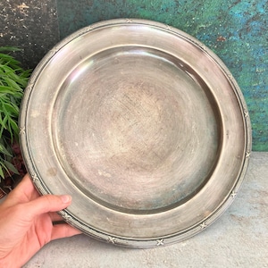 Peut inclure: Une assiette ronde argentée avec un large bord et une bordure décorative ornée de petits motifs en forme d'étoiles. L'assiette présente un aspect légèrement usé, suggérant qu'elle est vintage ou antique. Une main tient l'assiette, avec de la verdure en arrière-plan.